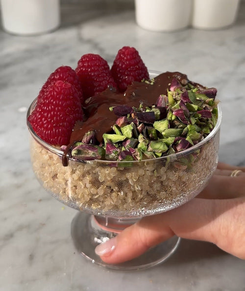 Dessert in a glass bowl with chocolate, pistachios, and raspberries held by a hand on a marble surface.
