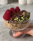 Dessert in a glass bowl with chocolate, pistachios, and raspberries held by a hand on a marble surface.