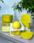 Yellow skincare product set with containers and a ball on a light surface with greenery in the background.