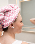 A woman wearing a pink and white patterned microfiber hair wrap turban headband, shown from the side with her reflection visible in a bathroom mirror.
