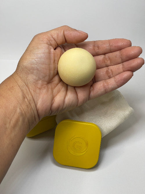 Hand holding a beige ball with a yellow container in the background