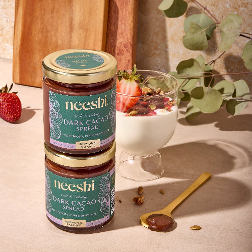 Neeshi dark cacao spread jars on a wooden surface with a dessert and spoon.
