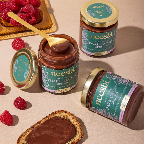 Jars of neeshi dark cacao spread with a spoon and raspberries on a beige surface