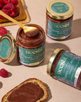 Jars of neeshi dark cacao spread with a spoon and raspberries on a beige surface
