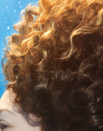A close-up of a person with voluminous curly hair in warm brown and golden blonde tones against a bright blue background.