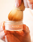 Hand holding a jar of NAPRIM naturals nourishing powder with a wooden brush.