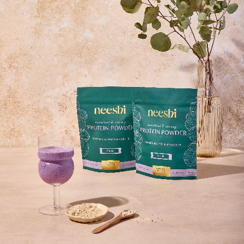 Two packages of neeshi protein powder with a glass of purple shake and a spoonful of powder on a neutral background.