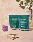 Two packages of neeshi protein powder with a glass of purple shake and a spoonful of powder on a neutral background.