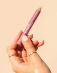 A hand holding a pink makeup pencil with the brand name 'Chellie' visible on the barrel, set against a solid beige background.