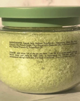 A clear plastic jar filled with pale yellow-green bath salts or scrub, topped with a muted green lid, photographed against a plain light background.