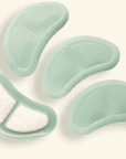 Four pale mint green adhesive pads or patches arranged on a light cream background, with one pad shown split open to reveal a white inner layer.