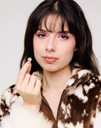 A close-up portrait of a woman with dark hair and pink eye makeup, wearing a brown and white cow-print faux fur zip-up jacket, holding a voltage skincare pimple patch between her fingers against a light gray background.