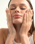 A close-up of a woman with her eyes closed, gently cleansing or applying skincare cream to her face with both hands. She is wearing a white ribbed tank top against a clean, bright, off-white background.