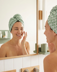 Woman applying skincare product in front of a mirror with a green towel on her head.