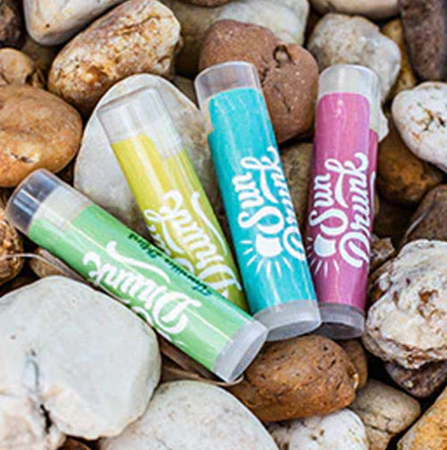 Colorful lip balms with 'Sun & Fun' branding on a bed of rocks.