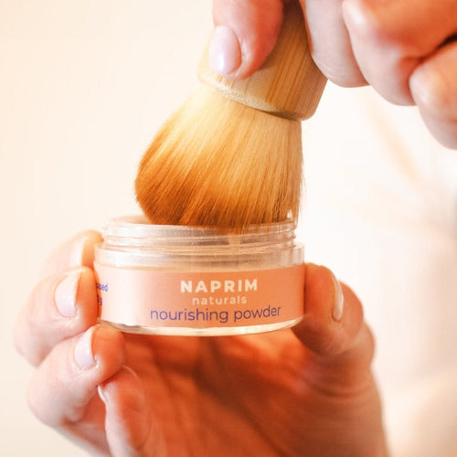 Hand holding a jar of NAPRIM naturals nourishing powder with a wooden brush.