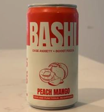 A Bashi Peach Mango beverage can with a red and off-white label, shown on a light surface against a neutral background.