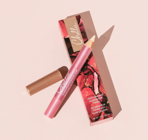 Chelio Illuminating Clay Crayon in a metallic pink tube with a gold tip, shown next to its floral pink packaging box and a separate brown clay stick applicator on a solid pale pink background.