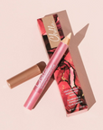 Chelio Illuminating Clay Crayon in a metallic pink tube with a gold tip, shown next to its floral pink packaging box and a separate brown clay stick applicator on a solid pale pink background.