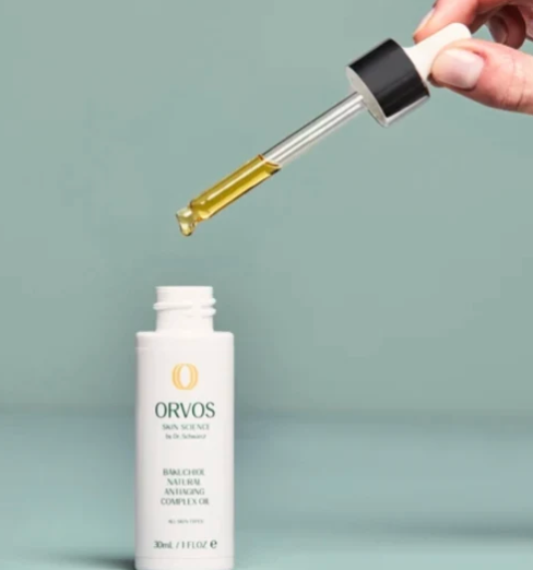 A hand holds a glass dropper above a white bottle of ORVOS Bio-Science Natural Antioxidant Complex Oil against a soft sage green background.