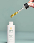 A hand holds a glass dropper above a white bottle of ORVOS Bio-Science Natural Antioxidant Complex Oil against a soft sage green background.