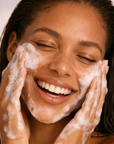 A close-up of a woman with her eyes closed, smiling while washing her face with foamy soap. Her hands and face are covered in white lather, and her dark hair is slightly damp against a soft light background.
