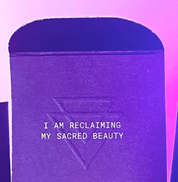 A close-up of a purple textured box with the embossed text 'I AM RECLAIMING MY SACRED BEAUTY' and a geometric symbol, under pink and purple lighting.