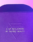 A close-up of a purple textured box with the embossed text 'I AM RECLAIMING MY SACRED BEAUTY' and a geometric symbol, under pink and purple lighting.