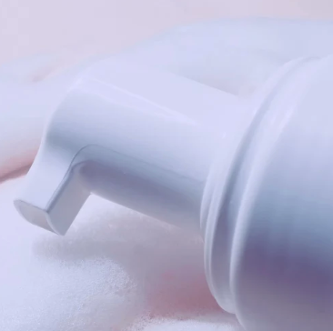 A close-up of a white pump dispenser dispensing a fine white foam against a soft pink and white background.