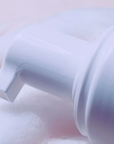 A close-up of a white pump dispenser dispensing a fine white foam against a soft pink and white background.