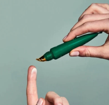 A close-up of a hand holding a green cosmetic tube with a gold applicator tip above another hand's index finger, against a muted sage green background.