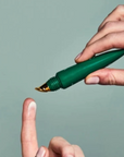 A close-up of a hand holding a green cosmetic tube with a gold applicator tip above another hand's index finger, against a muted sage green background.
