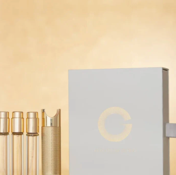 A luxury skincare or cosmetic set featuring a large white box with a gold circular 'G' logo, alongside three clear glass vials and a textured gold cap, set against a warm beige background.