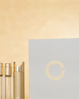 A luxury skincare or cosmetic set featuring a large white box with a gold circular 'G' logo, alongside three clear glass vials and a textured gold cap, set against a warm beige background.