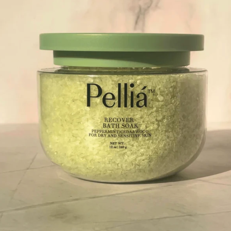 A clear plastic jar of Pellía Recover Bath Soak filled with pale yellow-green crystals, topped with a muted sage green lid, set against a soft marble background.