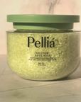 A clear plastic jar of Pellía Recover Bath Soak filled with pale yellow-green crystals, topped with a muted sage green lid, set against a soft marble background.