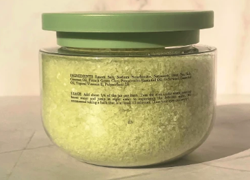 A clear plastic jar filled with pale yellow-green bath salts or scrub, topped with a muted green lid, photographed against a plain light background.