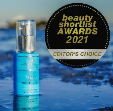 Viking Beauty Organic Sunscreen Night Cream bottle in a bright blue bottle with a silver pump, shown next to a black circular badge that reads 'beauty shortlist AWARDS 2021 EDITOR'S CHOICE'. The background is a blurred blue water scene.