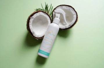 A white pump bottle of Coconut & Rosemary Hair Growth Serum by The Hair Brand, styled on a light green background with a halved coconut and a sprig of green leaves.