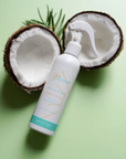 A white pump bottle of Coconut & Rosemary Hair Growth Serum by The Hair Brand, styled on a light green background with a halved coconut and a sprig of green leaves.