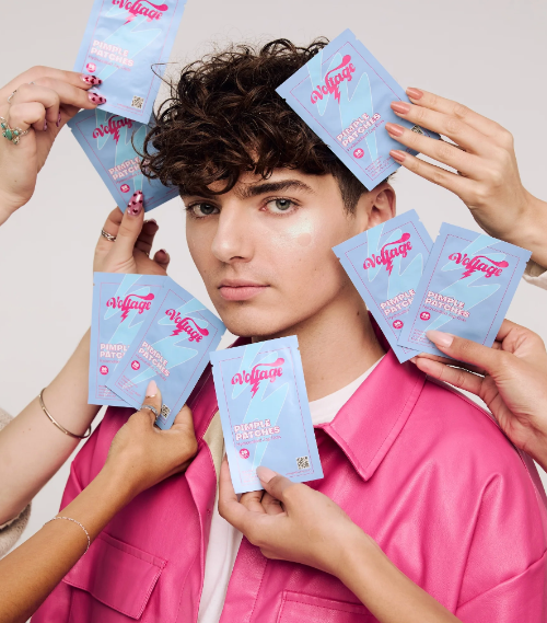 A person in a pink shirt surrounded by multiple hands holding light blue and pink Voltage skincare pimple patch packets against a plain light background.