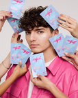 A person in a pink shirt surrounded by multiple hands holding light blue and pink Voltage skincare pimple patch packets against a plain light background.