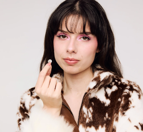 A close-up portrait of a woman with dark hair and pink eye makeup, wearing a brown and white cow-print faux fur zip-up jacket, holding a voltage skincare pimple patch between her fingers against a light gray background.