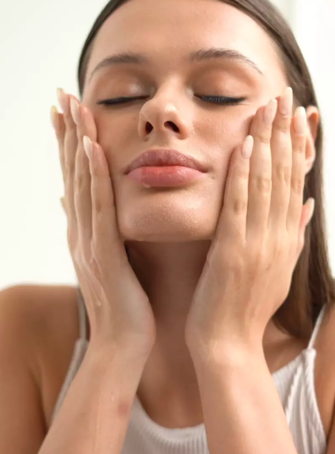 A close-up of a woman with her eyes closed, gently cleansing or applying skincare cream to her face with both hands. She is wearing a white ribbed tank top against a clean, bright, off-white background.
