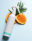 A bottle of Espieve Revive Body Spray with a white trigger sprayer, styled with sliced oranges and green palm leaves on a light blue background.