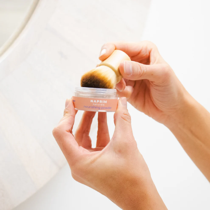 Person holding a makeup brush over a NAFIN product container on a light background