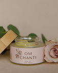 Jar of 'OM Chanti' product with a wooden lid and pink rose on a neutral background